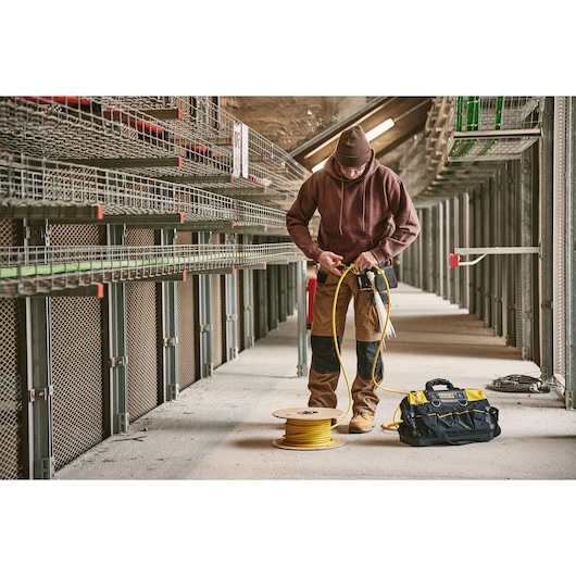 STANLEY 0-84-004 tool bag in yellow and black on floor near worker handling yellow cable spool in industrial setting.