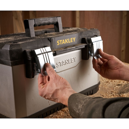 Photo of STANLEY 1-95-615 toolbox with hands opening metal latches, view from front in workshop setting.