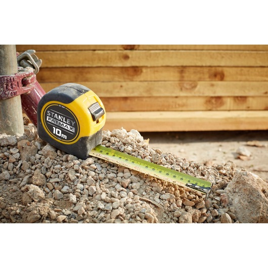 Stanley 33-829 tape measure with yellow casing on rocky ground, tape extended, wooden boards in background.