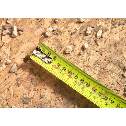 STANLEY 33-893 tape measure extended on a dusty plywood surface with small rocks scattered around, top view.