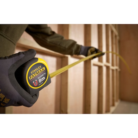 Stanley FMHT33869 tape measure with yellow blade extended, held by gloved hand at construction framing.