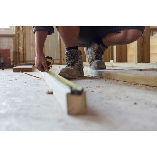 STANLEY FMHT36328 tape measure in use measuring wooden beam, person in work boots kneeling on construction site.