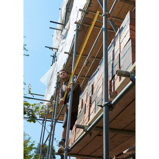 STANLEY FMHT36328 long yellow tape measure used by worker on construction scaffolding, bricks stacked nearby.