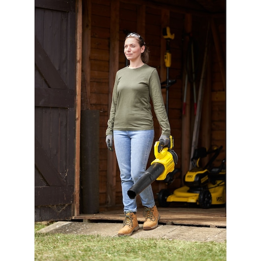 STANLEY SFMCBL730M1 leaf blower held by person outdoors, yellow body, shown in yard near wooden shed.