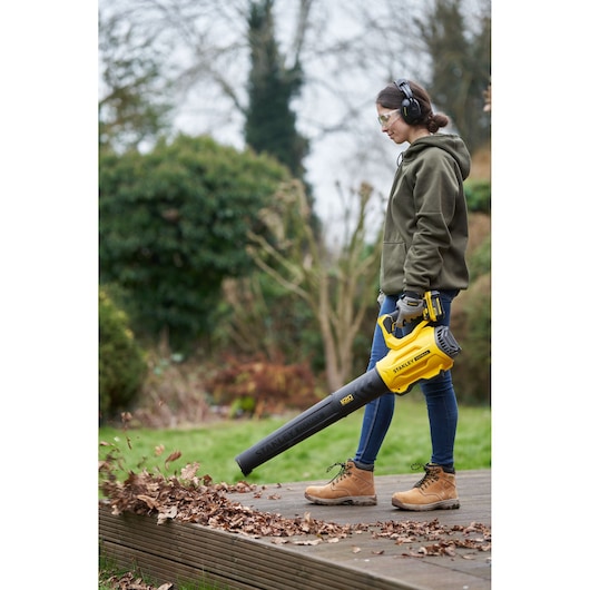 Photo of person using STANLEY SFMCBL7M1 cordless leaf blower clearing leaves from wooden deck outdoors.