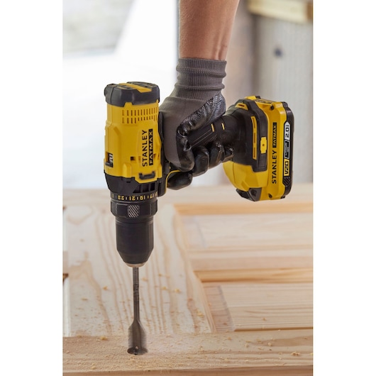 Photo of STANLEY SFMCD700D2S cordless drill driving a spade bit into wood, shown in use with a gloved hand.