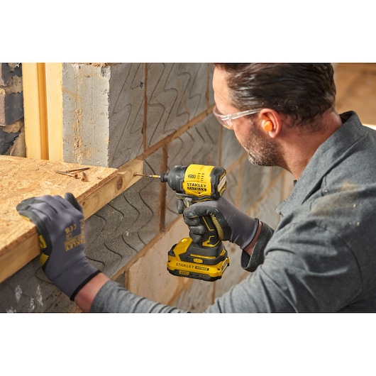 Photo of worker using STANLEY SFMCF811B-XJ cordless impact driver on wood near brick wall, wearing gloves.