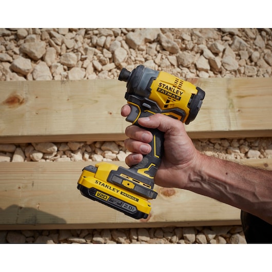 Stanley SFMCF820B impact driver held in hand, yellow and black, outdoor work site, side angle.