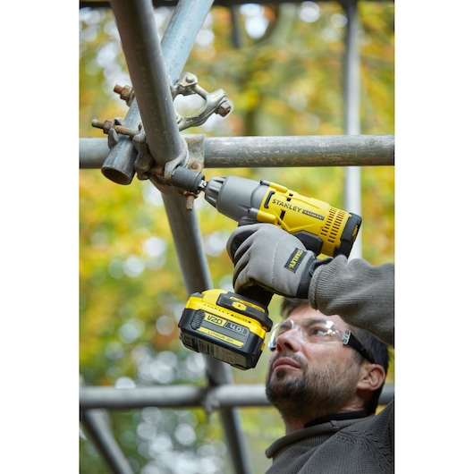 Photo of STANLEY SFMCF900B impact wrench fastening scaffold pipes, held by gloved hands outdoors.