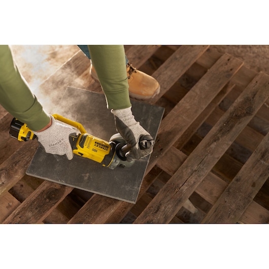 Photo of STANLEY SFMCG400B cordless angle grinder in use on a tile, person wearing gloves and work boots.