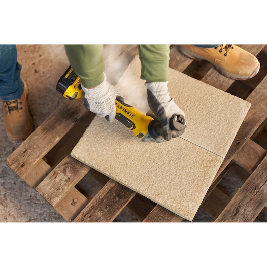 Photo of STANLEY SFMCG700B grinder cutting stone tile, held by gloved hands on wooden pallet.
