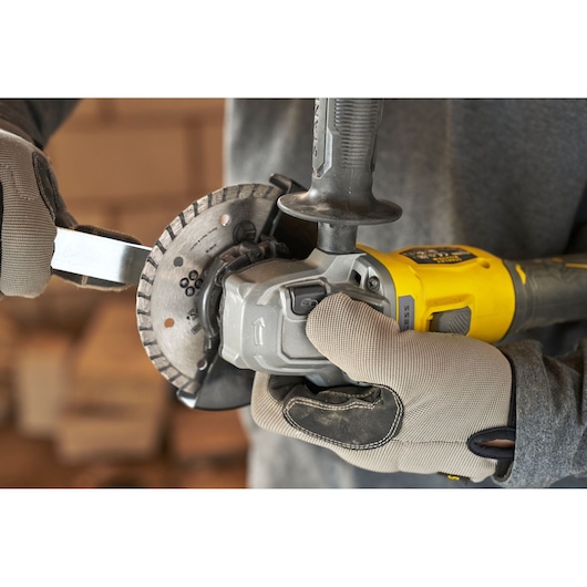 STANLEY SFMCG710B-XJ angle grinder, close-up side view, yellow and grey tool in use, gloved hands adjusting blade.