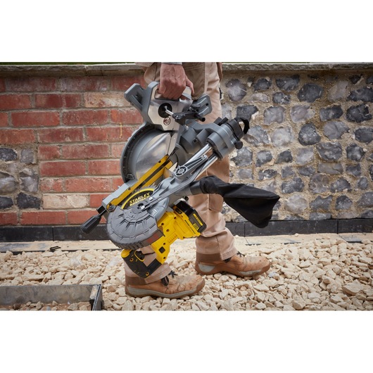 Photo of STANLEY SFMCS701B cordless miter saw carried by a person outdoors, yellow and gray, compact design.