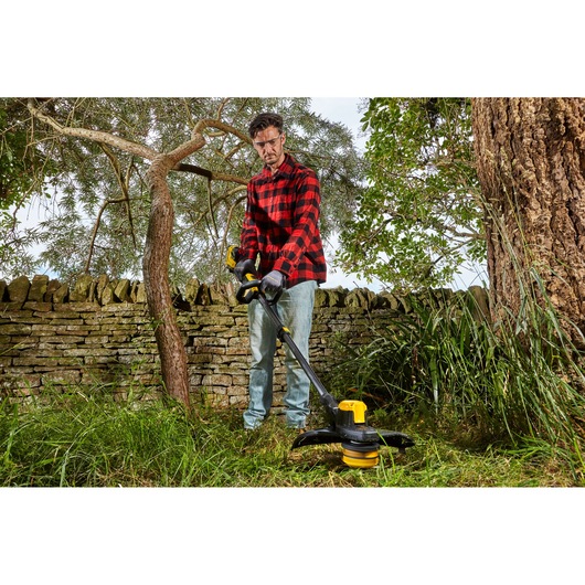 STANLEY SFMCSTB933M string trimmer used for grass trimming in a garden with stone wall and trees.