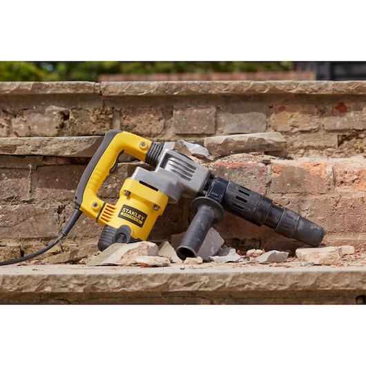 Photo of STANLEY SFMEH220K-QS demolition hammer resting on broken brick steps, yellow and black body, side handle visible.
