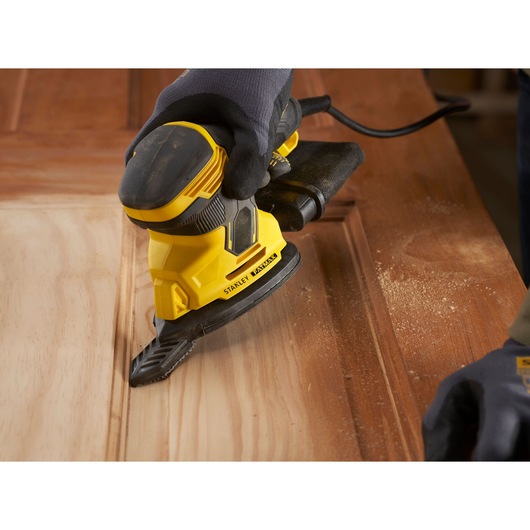 Photo of STANLEY SFMEW210S sander in use on wooden surface, close-up angle showing dust and ergonomic design.