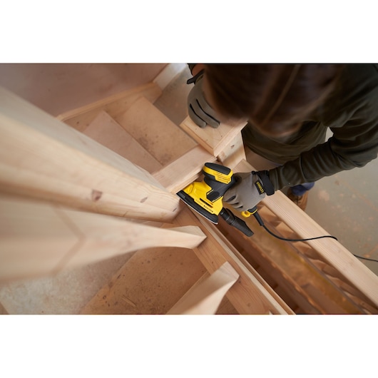 STANLEY SFMEW210S sander in use sanding wooden stairs, shown from above, with user wearing gloves.