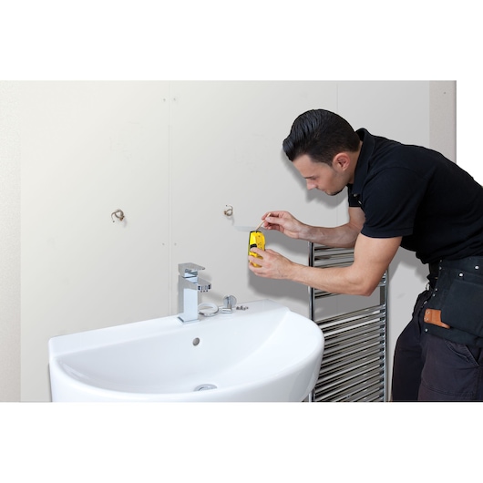 STANLEY STHT0-77403 stud finder in use above bathroom sink, worker in black shirt holding tool against wall.