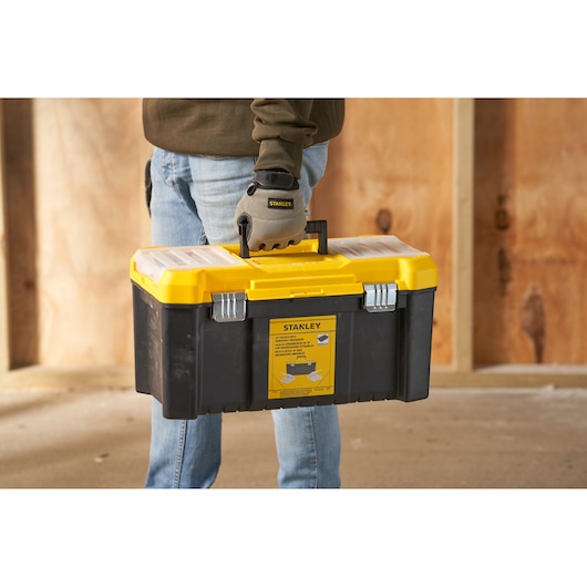 Photo of STANLEY STST75787-1 tool box with yellow lid, black base, carried by a gloved hand, front angle.
